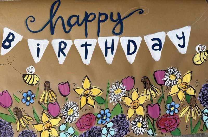 Hand-painted happy birthday banner on kraft paper with navy script 'happy' above white pennant bunting spelling 'BIRTHDAY,' and a flower garden of daffodils, tulips, roses, daisies, forget-me-nots, and buzzing bees